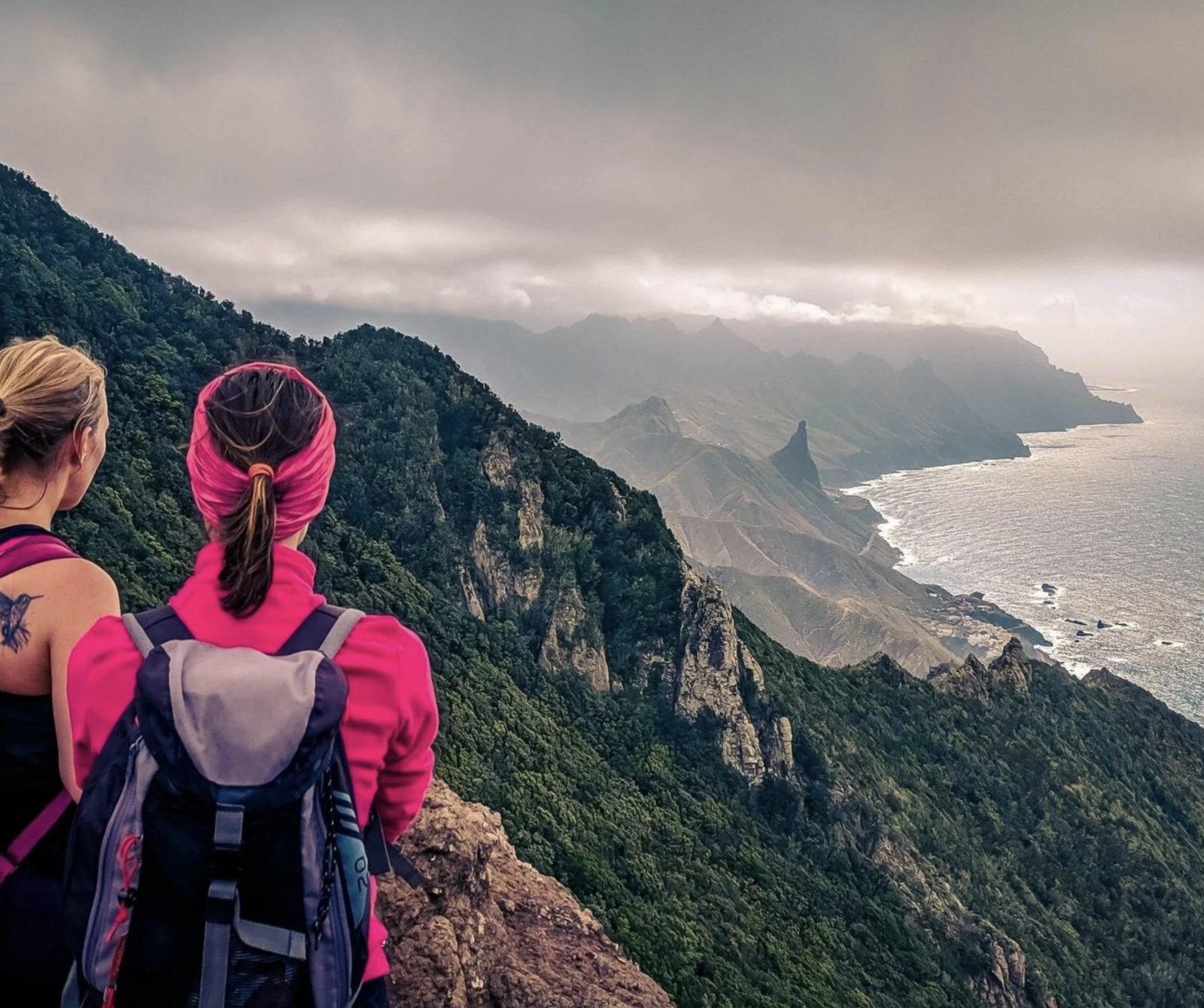 Two women with backpacks looking out over green mountains and the Atlantic coast